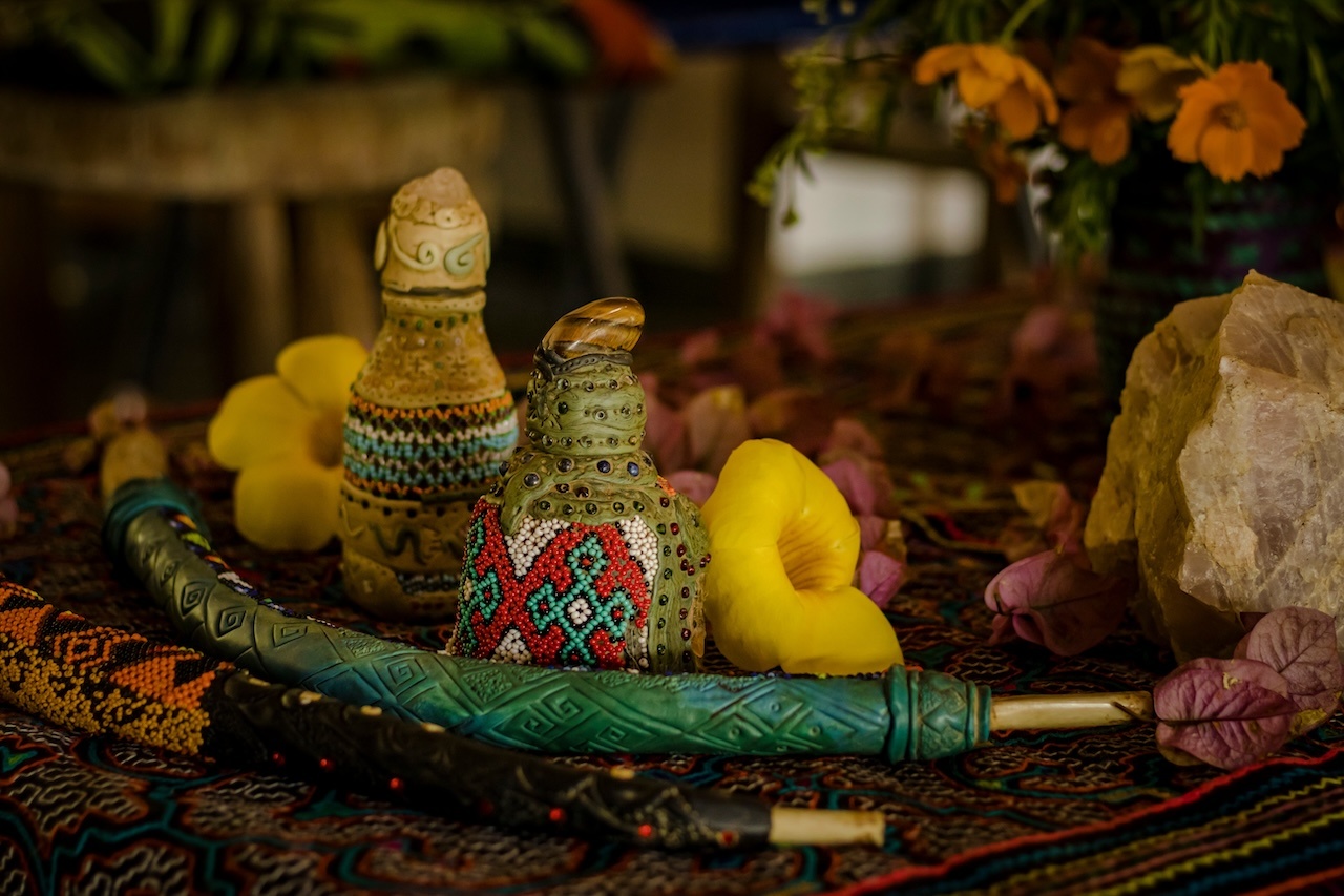 Traditionelles Rapé-Set mit verzierten Fläschchen, Tepi-Pfeife, Blumen und Kristall auf einem bunten gewebten Tuch, verwendet in schamanischen Ritualen.