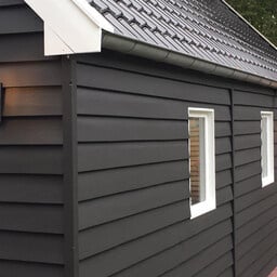 Detail einer matt schwarz gestrichenen Holzfassade mit weiß abgesetzten Fenstern und warmem Holzakzent