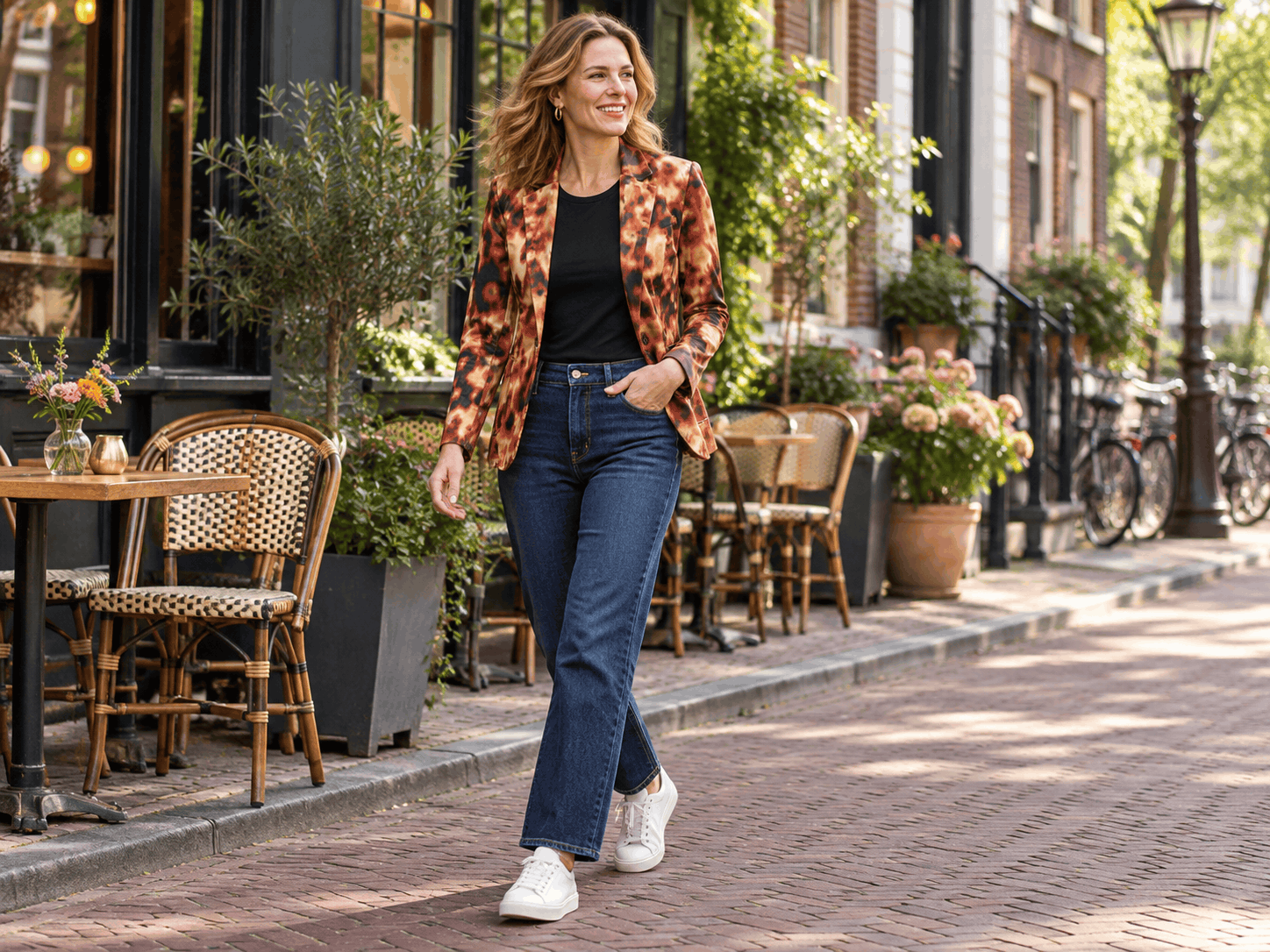 Een glimlachende vrouw die over een Amsterdamse straat loopt in een panterprint blazer, een zwart shirt, donkere jeans en witte sneakers.