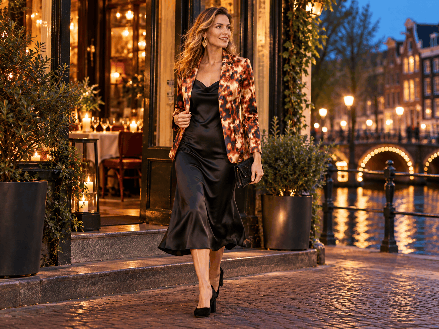 Een vrouw die 's avonds langs een Amsterdamse gracht loopt in een panterprint blazer, een zwarte jurk en hakken, met een clutch in haar hand.