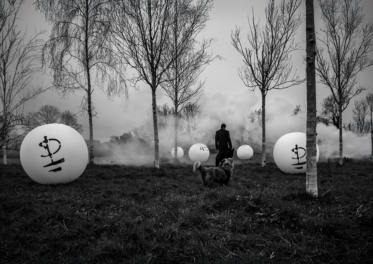 Black and white photograph of a figure and dog among birch trees, white spheres and smoke, by Polly Samson