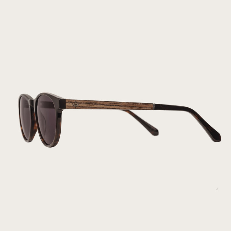 La paire de lunettes l’Ellipse ‘Forever Havanas Black’ poséde un cadre en écaille marron foncé avec des verres noirs. Fabriquée à partir de bois durable et du bioacétate Mazzuchelli Italien avec des branches et écaille de tortue en ébène et manchons en ac