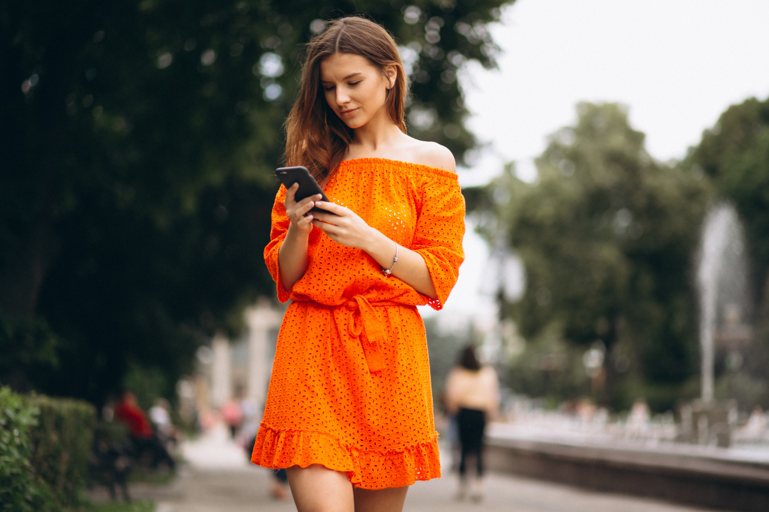 vrouw met smartphone loopt op straat in een oranje zomerjurkje.