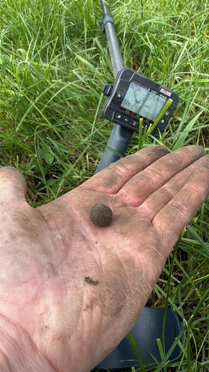 Net opgegraven musketkogel in een hand vol aarde, met de metaaldetector op de achtergrond in het gras