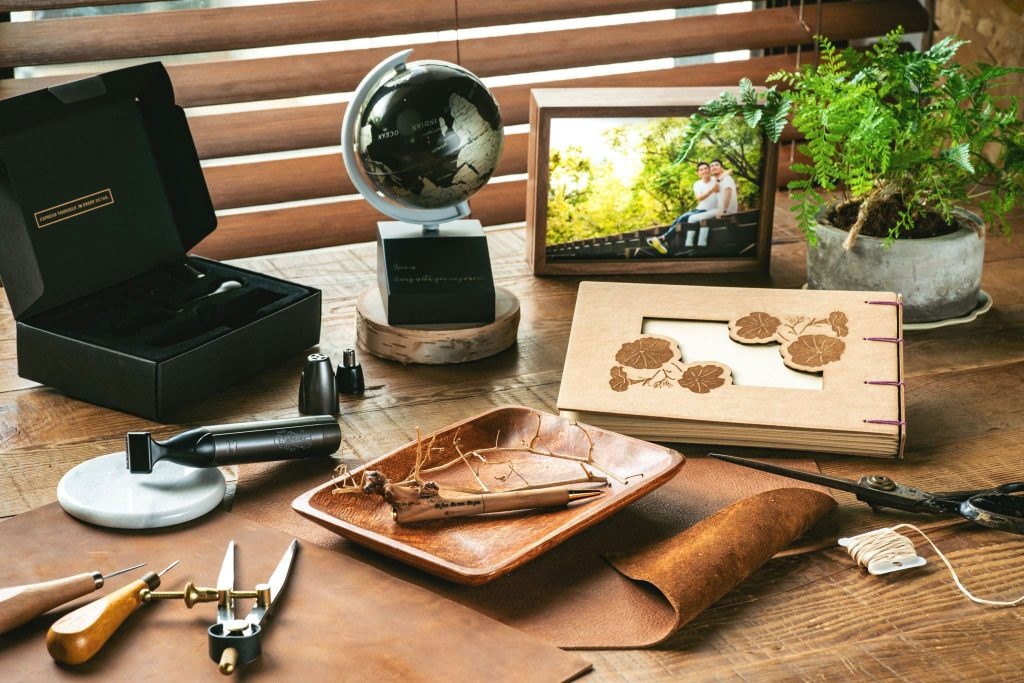 A desk filled with engrave and cut examples: A metal reasor engraved, a felt box with engraving on, wooden book cover with an engraving of some flowers and a hole where you can see the first page, an engraved wooden pen and a pease of leader ready to be engraved or cut.