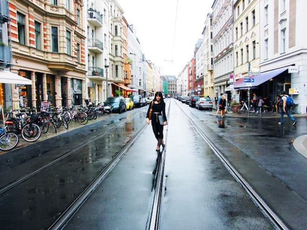 Eine Frau, die auf einer nassen Straße in einer städtischen Umgebung in der Nähe der Brewing Beauty Company in Berlin Mitte mit Gebäuden und geparkten Fahrrädern über Straßenbahnschienen läuft