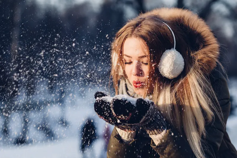 A woman in a winter jacket and earmuffs blows snow from her hands in a snowy landscape, capturing a joyful, wintry moment that reminds us of seasonal skin care needs.