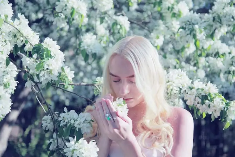 A woman with blonde hair gently smelling white blossoms on a sunlit bush, surrounded by abundant flowers, evoking a serene summer atmosphere.