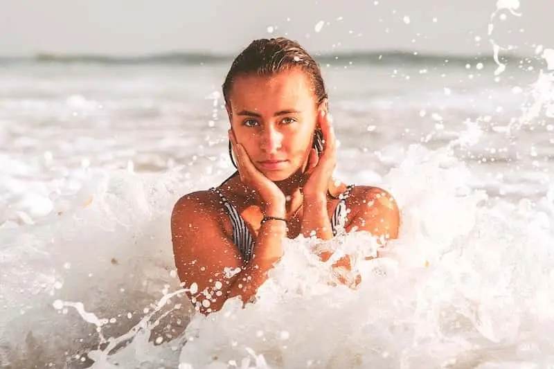 A young woman in the ocean with her hands on her cheeks, splashed by foamy waves, looking directly at the camera, demonstrating summer skin care.