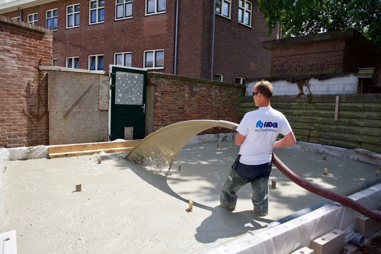 Een man die de betonvloer stort. Hij staat met zn laarzen in het beton met een shirt waar betonpompen op staat. Wat rode huisjes op de achtergrond.