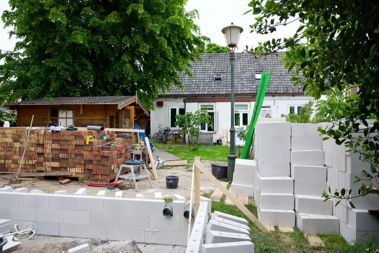 kalkzandsteenblokken op elkaar gestapeld met metselstenen ernaast. Een klein wit huisje op de achtergrond.
