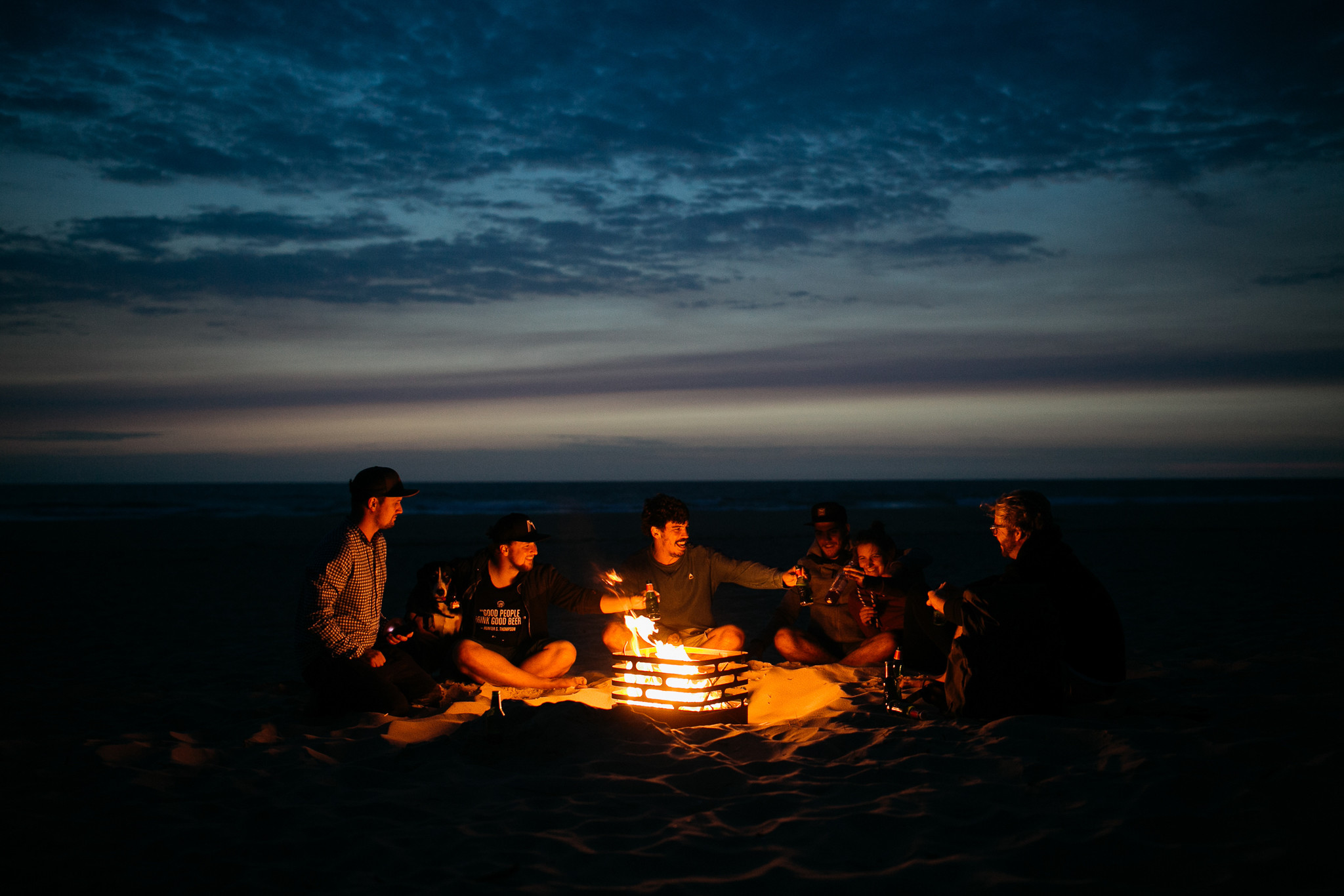 Vriendengroep jonge mensen met bier rond Hofats vuurkorf op het strand