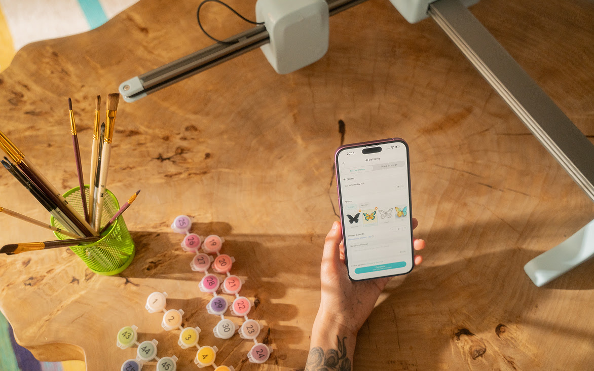 Mano che tiene un cellulare con l'applicazione LOKLiK IdeaStudio aperta su uno sfondo di tavolo in legno. Sul tavolo ci sono una LOKLiK iPaint e i colori del set di vernici.
