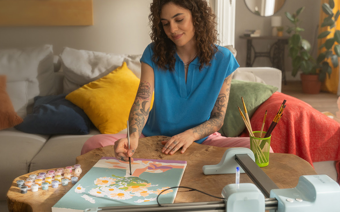 Donna a un tavolo basso che dipinge un vaso con fiori su una tela. Sul tavolo c'è un LOKLiK iPaint