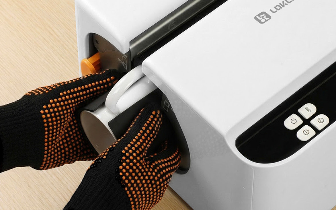 Close-up view of a person wearing heat-resistant gloves loading a white ceramic mug into the automated slot of the LOKLiK Auto Mug Heat Press.