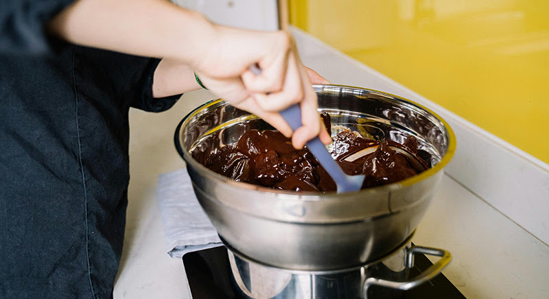 Hand holding a spatula stirring chocolate on a double boiler against a yellow background
