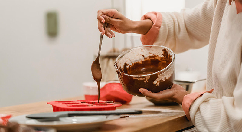 Woman holding a spoon and pouring melted chocolate in a red sillicone mould.