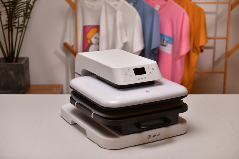 Central on a empty table stands the LOKLiK Auto Heat Press LOKLiK White with behind the table colored T-shirts with each a different design on them on coathangers.