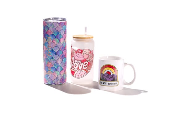 Stainless steel tumbler with sublimated colourful shell motif, glass mug with lid and straw and sublimated heart shape and love text, sublimated ceramic mug with red and yellow rainbow, mountain range and 'the most beautiful' in text.