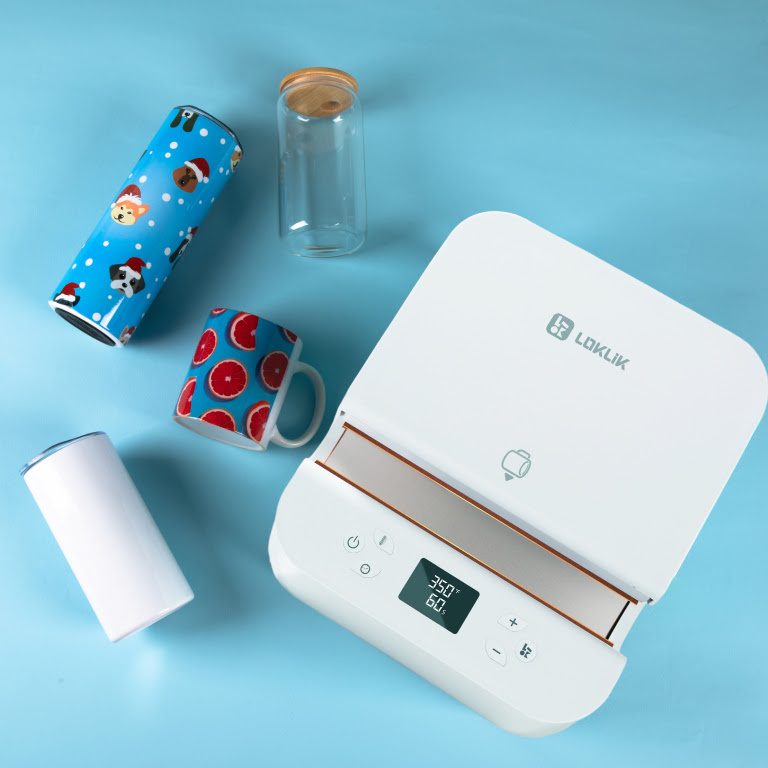 Top down view of an Auto Tumbler Heat Press on a light blue table. Text ot it are two tumblers, a mug and a jar. The timbler and mug have a sublimation print.