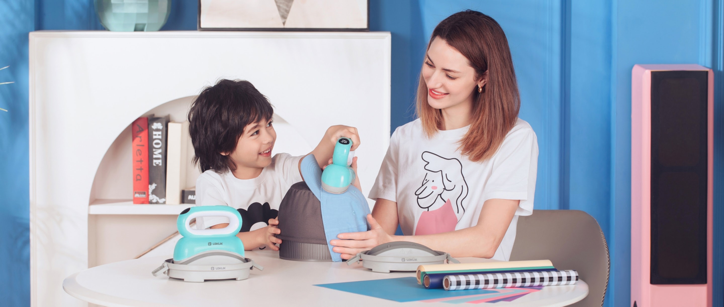 A woman and child sitting ata round table. The child is holding the LOKLiK Impress™ Hat and is ironing a design on a light blue hat. There is an extra LOKLiK Impress™ Hat standing on the table and several rolls of heat transfer vinyl.