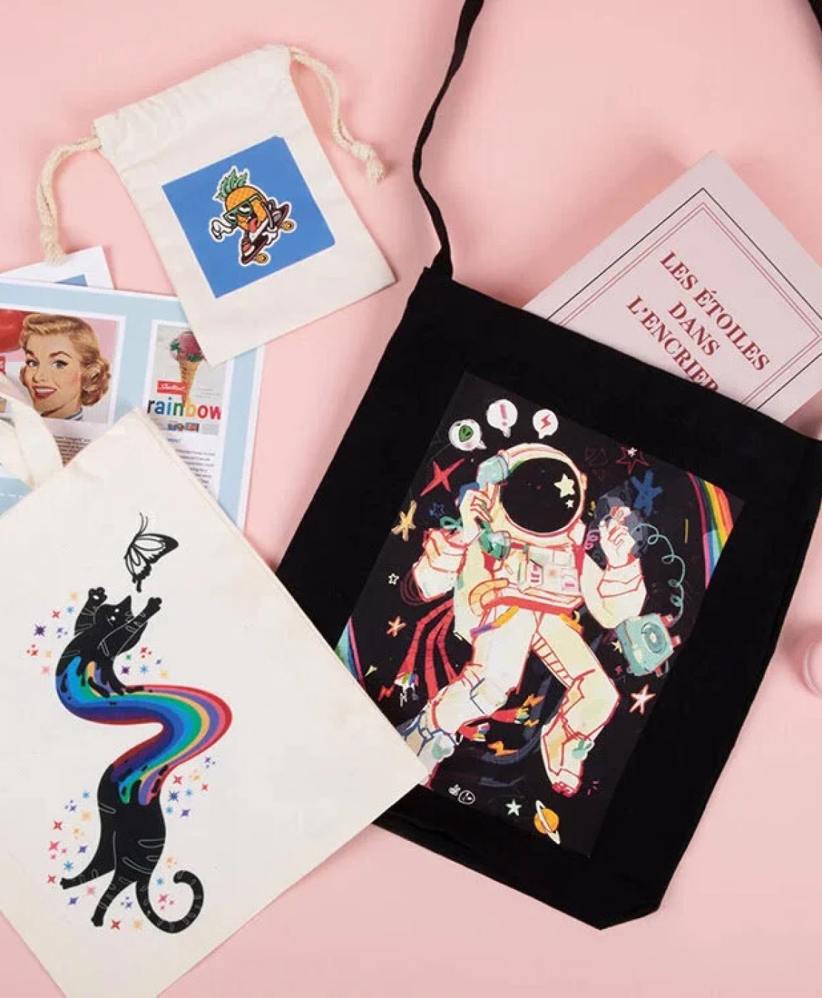 Top down view of a white and a black tote bag on a light pink background. The black one has a heat transfer design of an astronaut. The white tote features a black cat chasing after a butterfly. Its body is made of a rainbow.