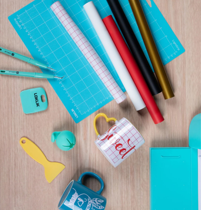 To down view of a wooden table with several rolls of adhesive vinyl and one roll of transfer tape on top of a blue cutting mat. On the left are weeding tools, a scraper and scrap collector. There are two mugs with a permanent adhesive vinyl design.