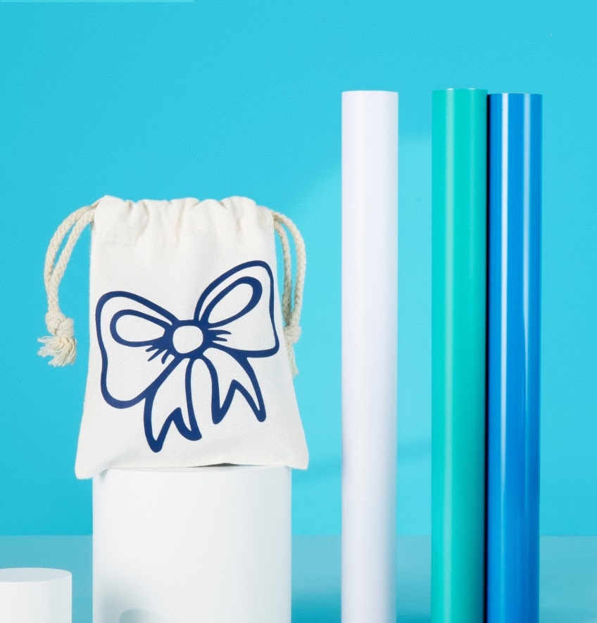 Three rolls of heat transfer vinyl in white, aqua blue and ocean blue. On the left is a small drawstring bag with an iron-on design of an outlined bow in blue vinyl.