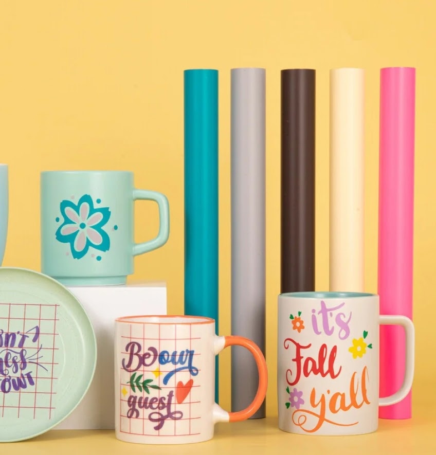 Five rolls of permanent adhesive vinyl in matte tiffany blue, light gray, dark coffee, matte beige and pink in front of a creamy yellow background. They are standing upright behind three mugs and a plate with different desings in permanent adhesive vinyl.
