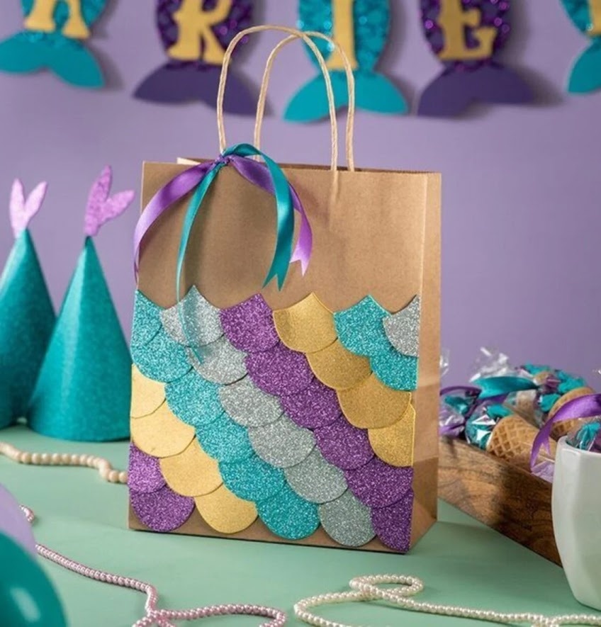 Paper bag decorated with a ribbon and a pattern forming scales made from glitter cardstock. In the background there are party hats and decorations.