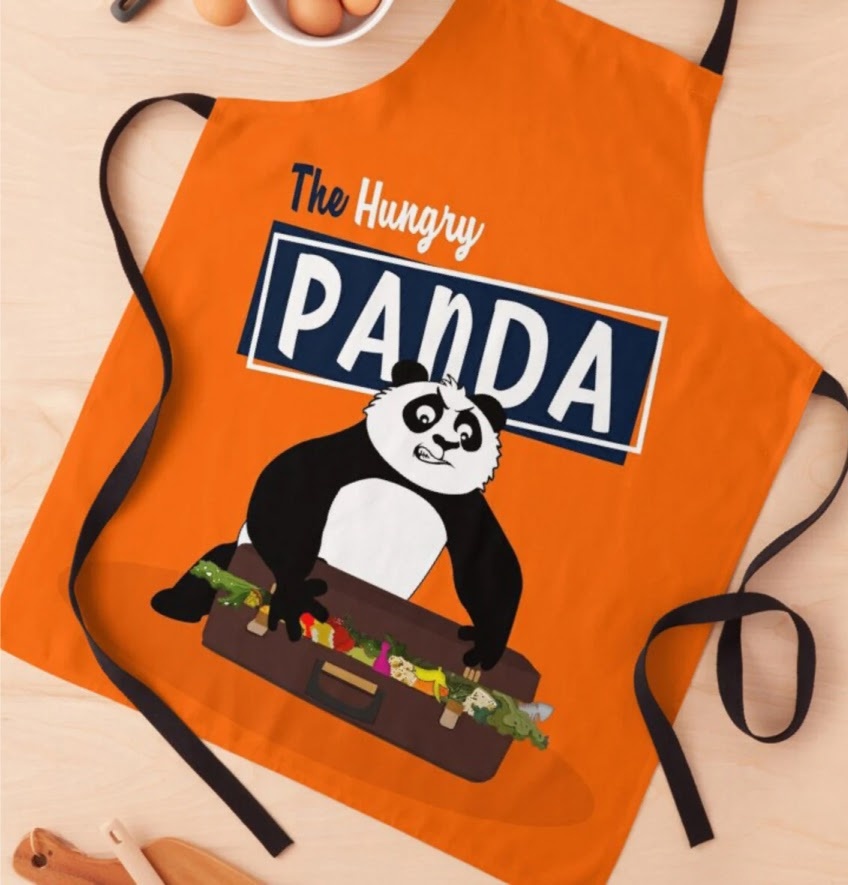 Top down view of an orange apron with a sublimation print of a cartoon panda stuffing food into a suitcase and the text 'The Hungry Panda'
