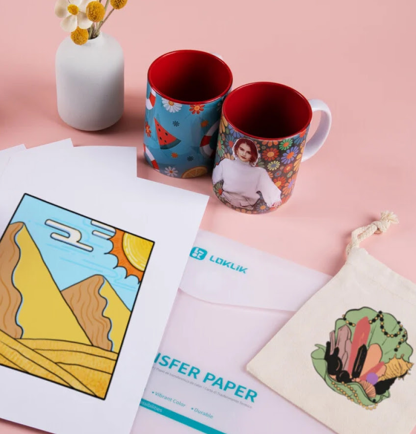 A sheet of heat transfer paper printed with a stylised design of mountains in a sunny landscape. It is placed on top of the envelope the sheets of heat transfer paper come in. Next to it is a canvas bag with a heat transfer print of gemstones and two mugs with heat transfer prints.