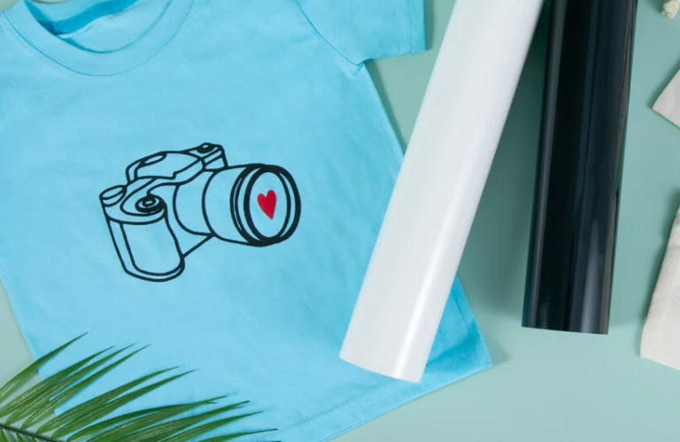 Top down view of a blue table with a light blue T-shirt with an iron-on design of a camera in black and red heat transfer vinyl. Next to it are two large rolls of heat transfer vinyl in white and black.