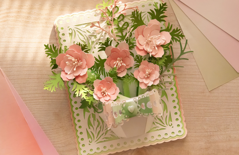 Top down view of a wooden table with an intricately cut paper card of a vase filled with pink flowers and green foliage. Next to it are sheets of paper.