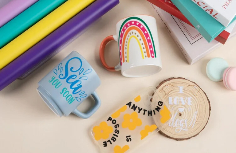 Top down view of a table with customised mugs and a phone case next to some rolls of permanent adhesive vinyl. One mugs says 'Sea you soon' in swirly letters, the other has a rainbow. The phone case says 'Anything is possible' in black vinyl surrounded by large flowers in orange vinyl.