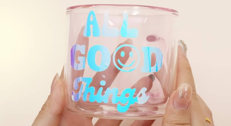 Twee handen houden een glazen beker vast met een ontwerp in holografische zelfklevende vinyl met de tekst 'ALL GOOD Things'.