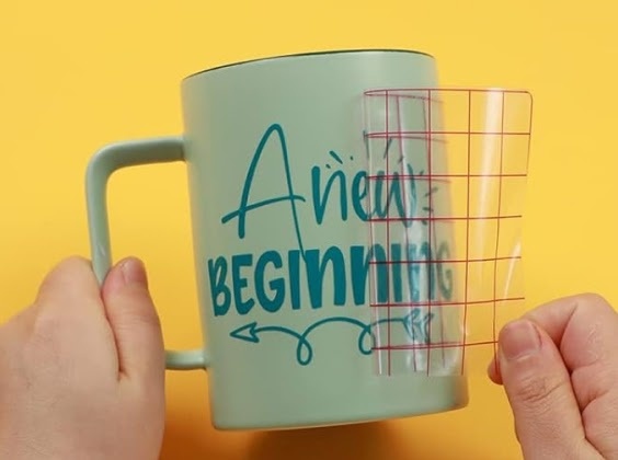Grid transfer tape transferring a lettering design in teal permanent adhesive vinyl on a mug. It says 'A new beginning'