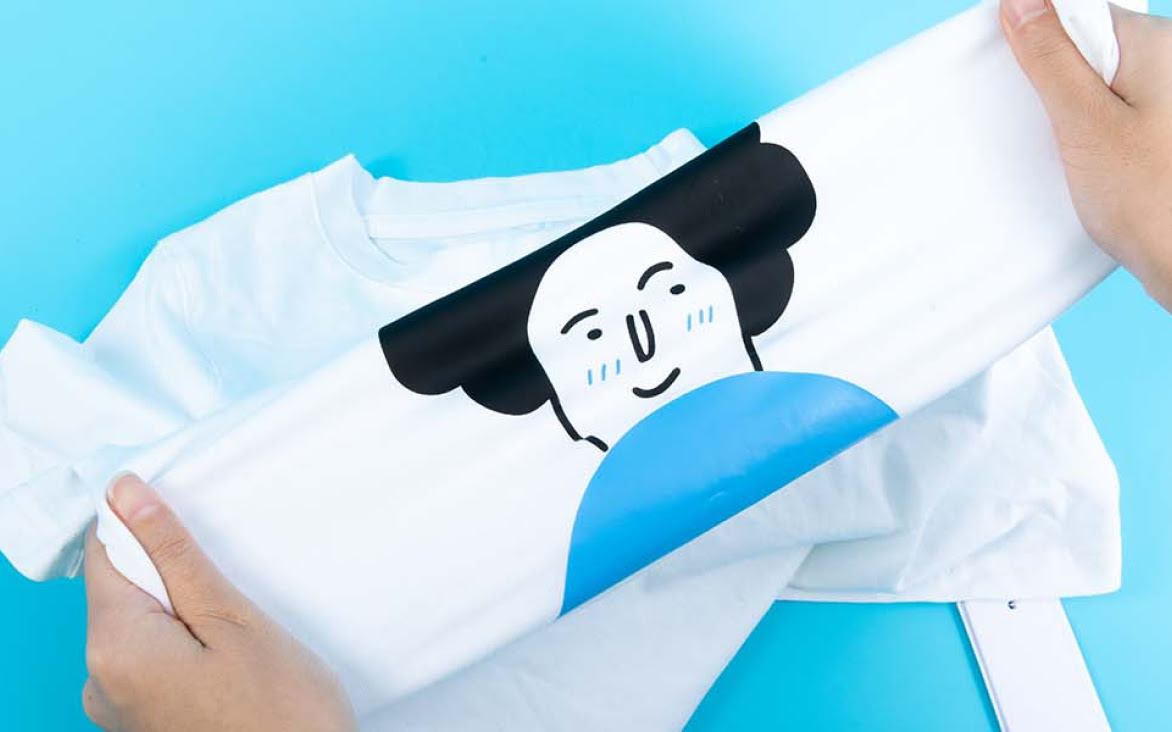 Twee handen die de stof van een wit shirt met een ontwerp van een geïllustreerd gezicht in flexfolie uitrekken.
