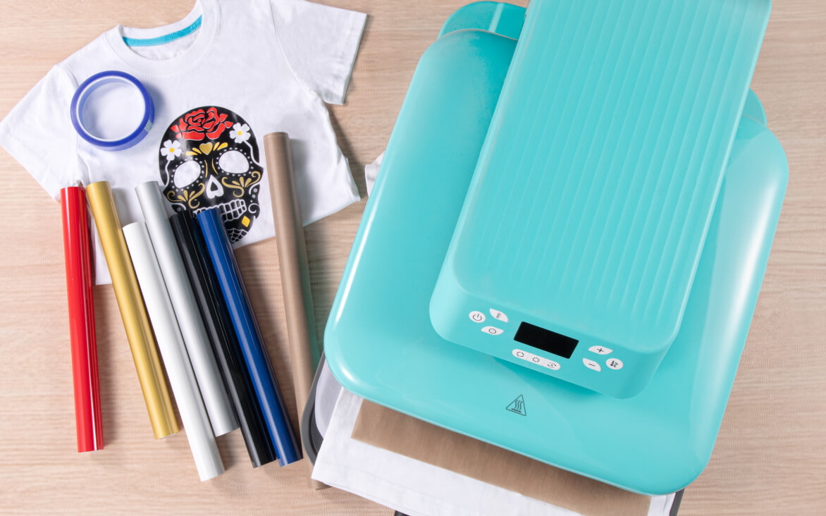 Top down view of a wooden table with a LOKLiK Auto Heat Press in LOKLiK Blue. On its left side is a white T-shirt with a iron-on mexican skull design. There is a roll of sublimation heat tape, 6 rolls of HTV and a roll of Teflon sheet.