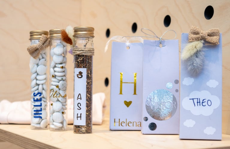 Baby shower favours on a wooden shelf. There are three plastic tubes filled with chocolates or tea and three small cardboard bags. All of these are decorated with the baby's name: Jilles, Olivia, Ash, Helena, Victor, Theo.