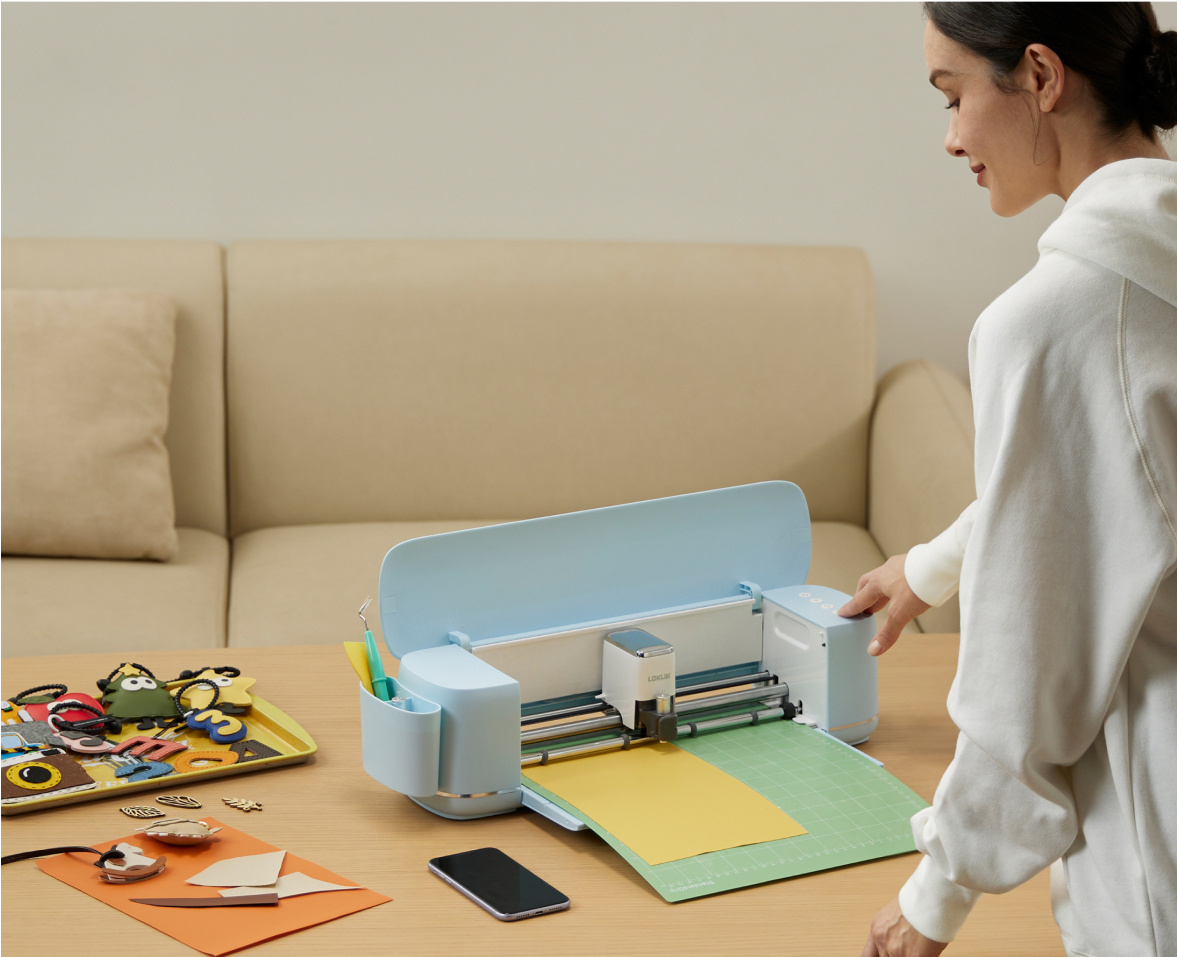 Frau drückt den Startknopf an einer LOKLiK iCraft™ in Pastellblau mit einer grünen Schneidematte und gelbem Material. Das Plotter steht auf einem Holztisch. Daneben liegen mehrere ausgeschnittene Formen aus verschiedenen Materialien.