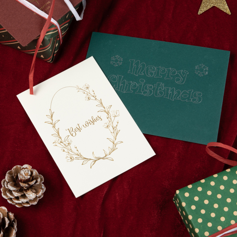 Dark green greeting card with the message 'Merry Christmas' in silver foil and a white card with a flower wreath and 'Best Wishes' in gold foil on a red table cloth surrounded by two gifts and some pinecones.