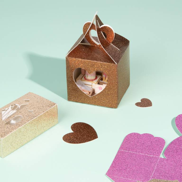 Folded box in orange glitter cardstock on a mint coloured table. The box has a heart shape cut out of one side and is filled with a small papercraft scenery. The box is surrounded by two hearts in orange glitter cardstock and one unfolded box. On the left is another small folded box.