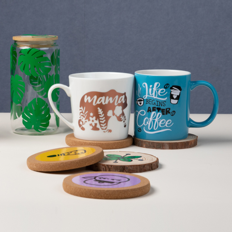 A white mug with a bear and the words mama in permanent adhesive vinyl and a blue mug with a black and white design  with the words 'Life begins after coffee'in permanent adhesive vinyl on two wooden coasters. On the left is a glass tumbler with a pattern of green palm leaves in permanent adhesive vinyl. In the foreground are three wooden coasters with vinyl designs.