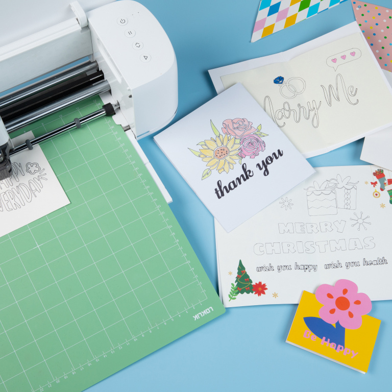 Top view of a LOKLiK iCraft™ in Daisy White on a light blue table with a green cutting mat loaded. On the right are several cards with desings made with the LOKLiK pens and cutting machine.