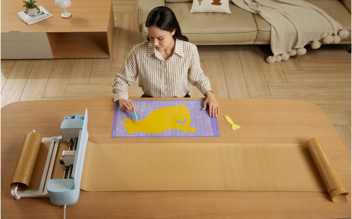 Woman sitting on a low table. She is weeding details from a cat in glitter vinyl on a purple cutting mat. In front of her there is a LOKLiK iCraft™ in Bubble Blue and the LOKLiK roll holder.