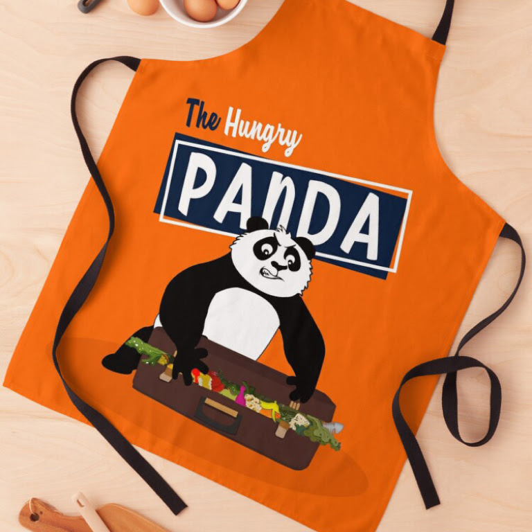 Top down view of an orange apron with a heat transfer design of a panda stuuffing food in a suitcase and the words The Hungry Panda.