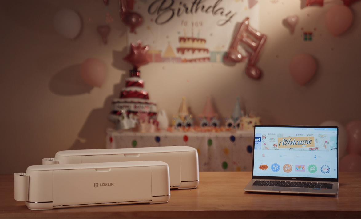 Two LOKLiK iCraft™ cutting machines in Daisy White on a wooden table. On the right there is an open laptop showing LOKLiK IdeaStudio™. In the background a wall and table are decorated for a birthday party: banners, hats, decorations.