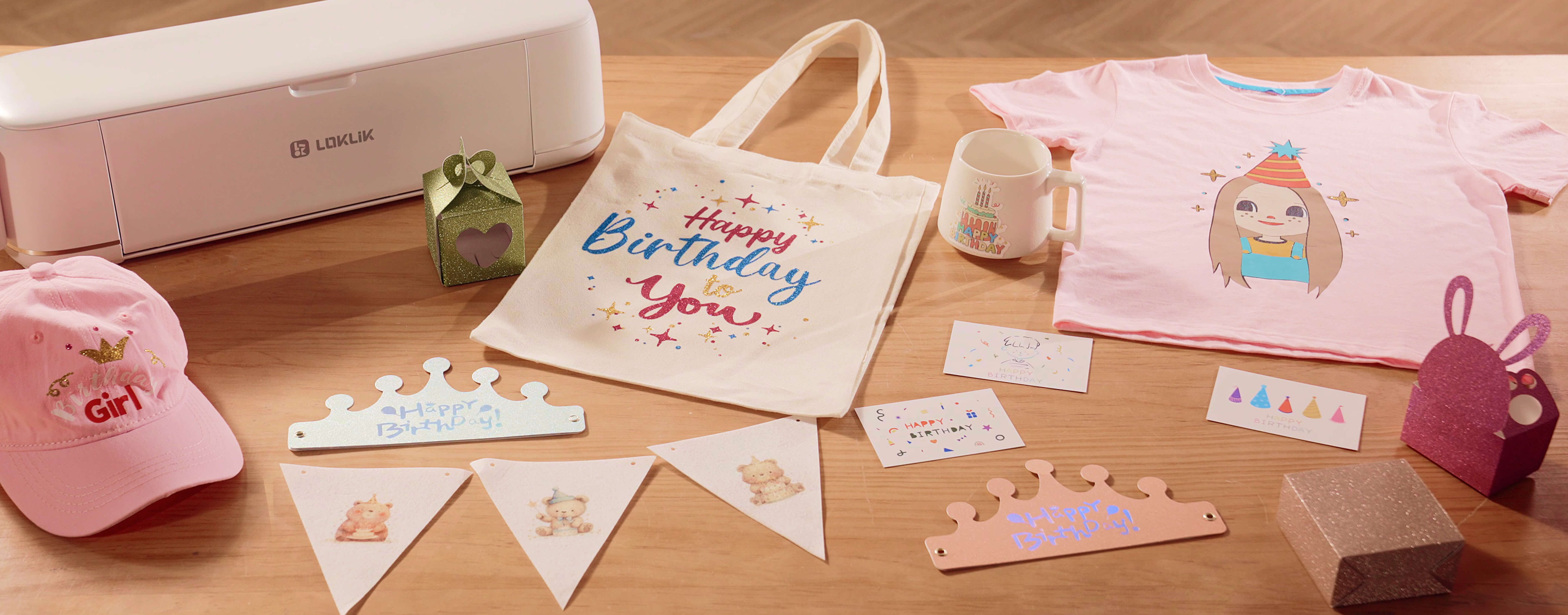 Wooden table with a LOKLiK iCraft™ in Daisy White. The table shows several projects made with the machine: a pink cap with an iron-on design saying 'Birthday Girl', two cut-out paper crowns with a foil design of 'Happy Birtday!', three folded boxes in glitter cardstock, a canvas tote with an iron on design in glitter heat transfer vinyl in pink, blue and gold saying 'Happy birthday to you', a mug with a custom sticker of a birthday cake, a pink T-shirt with an iron-on design of a girl in a birthday hat, three small flags with little bears printed on heat transfer paper and ironed on and three smaller cards with a design drawn on.
