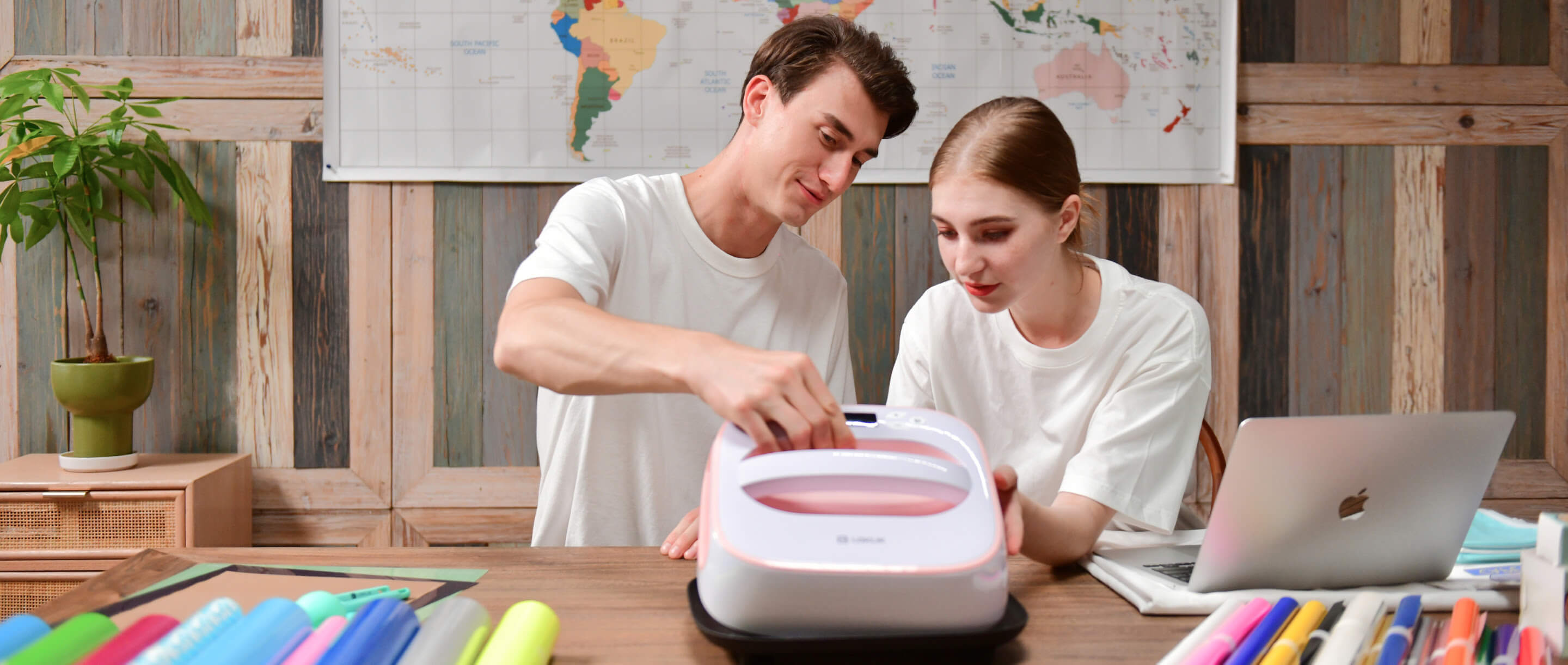 A man and a woman sitting at a table next to each other. The are holding a LOKLiK Easy Heat Press in Romance Pink and raising it from it safety base at an angle. There is also an open latop on the table and several rolls of heat transfer vinyl in various colours. In the background there is a map of the world on the wall.